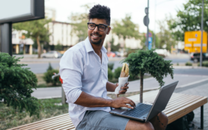 Young man eating a sandwich on a bench outside with his laptop on his lap