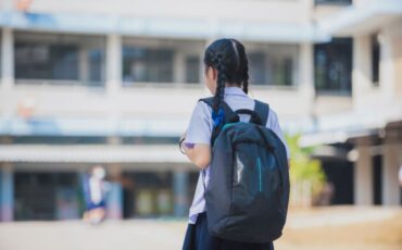 Image of young kid facing school, wearing her backpack