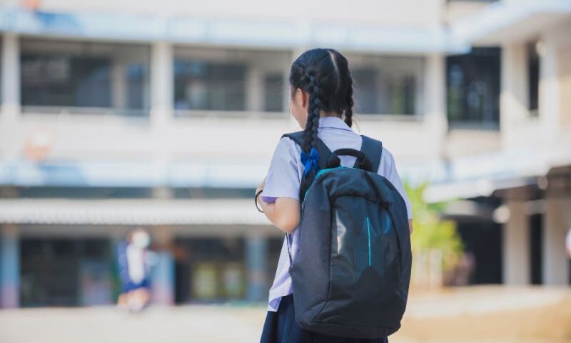 Image of young kid facing school, wearing her backpack