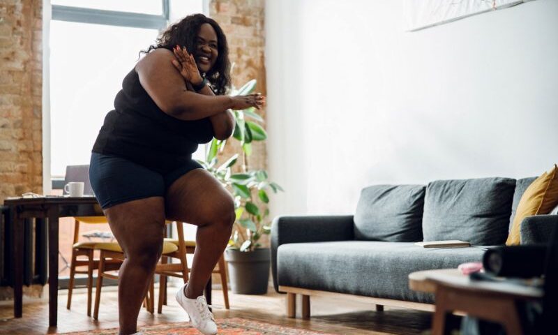 Image: Young black woman affected by obesity does a workout inside her living room during the day