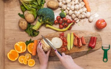 Image of someone's hands as they meal prep by cutting produce on a cutting board. In the image are oranges, broccoli, peppers, pickles, garlic and other ingredients.