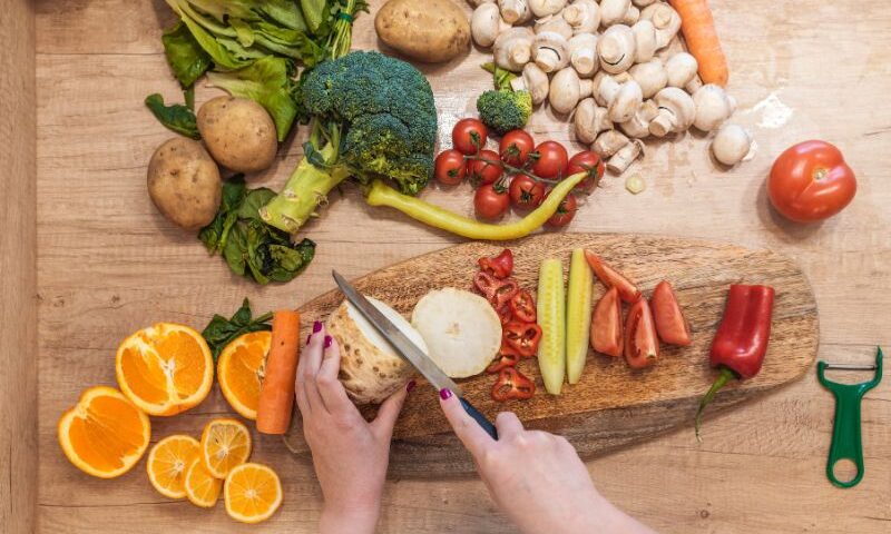 Image of someone's hands as they meal prep by cutting produce on a cutting board. In the image are oranges, broccoli, peppers, pickles, garlic and other ingredients.