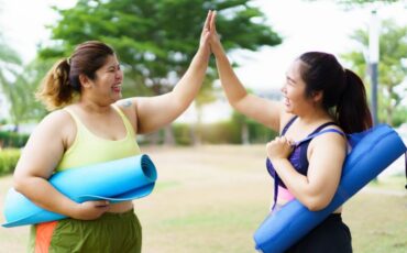 Two women high five in a park, holding their yoga mats